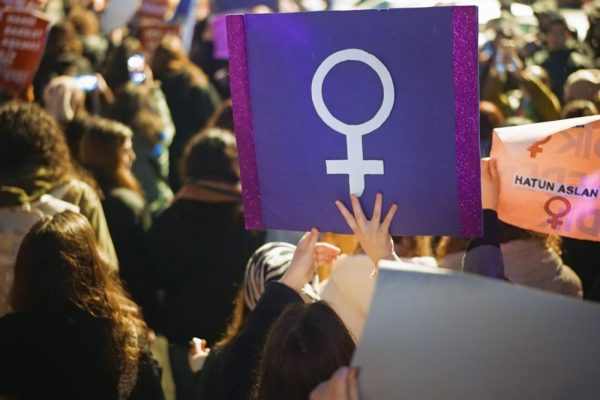 Article Banner - Women protest holding sign with female symbol in white against a purple background